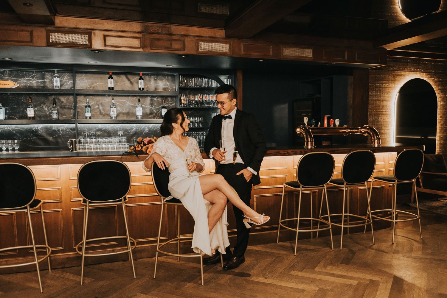 Elegant couple in formal attire sits at an upscale bar with warm lighting and leather barstools.
