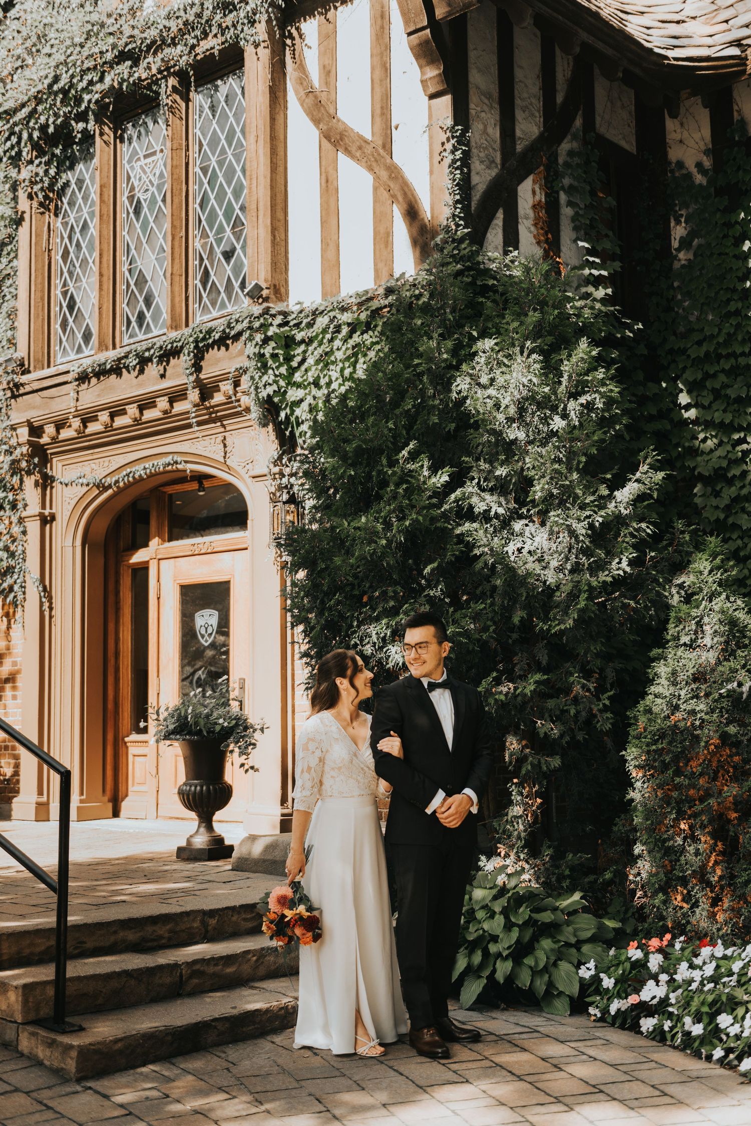 Elegant wedding couple poses in front of historic Tudor-style mansion with manicured garden and stone pathway.