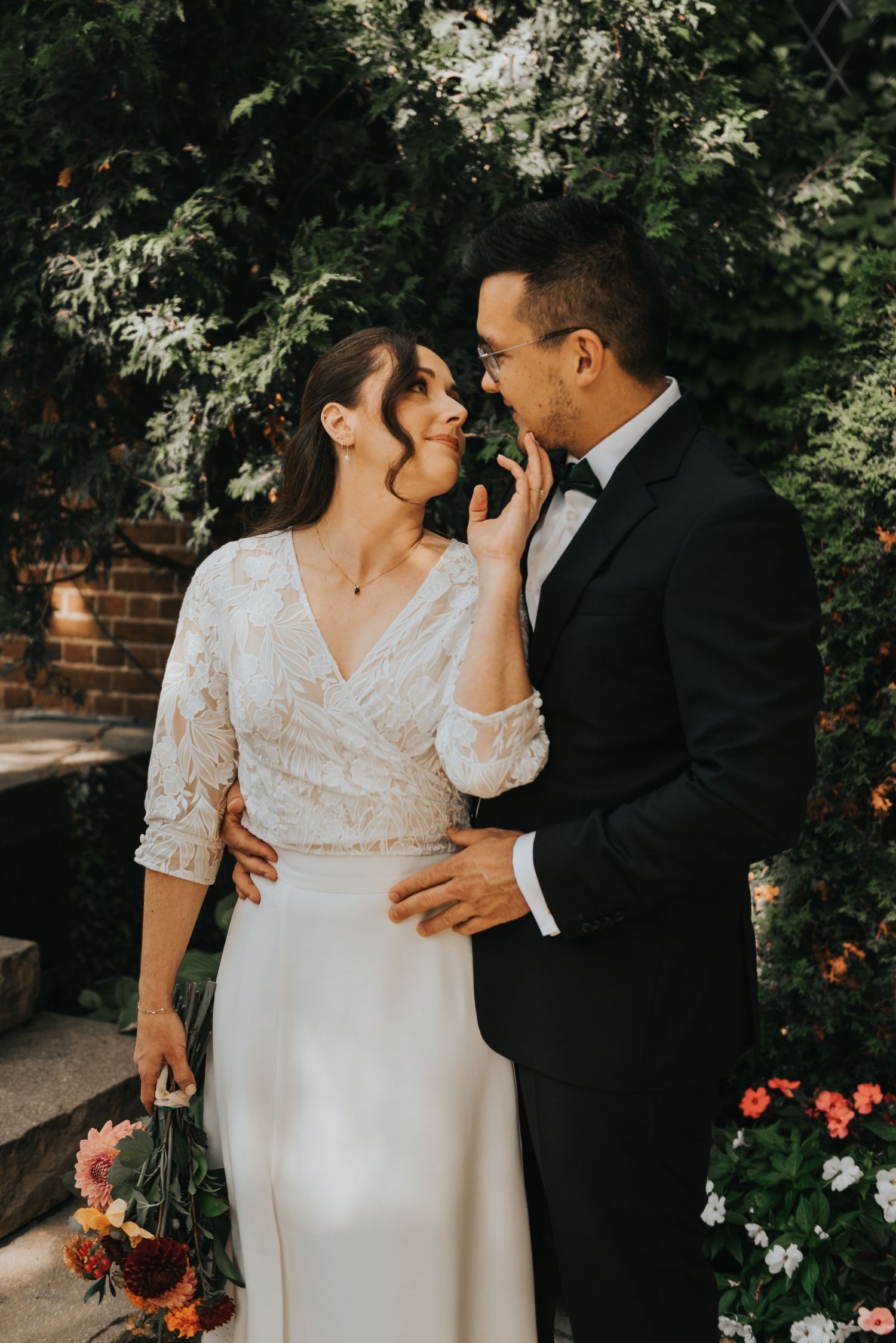 A romantic wedding couple embraces in a garden setting with lush greenery and flowers during golden hour.