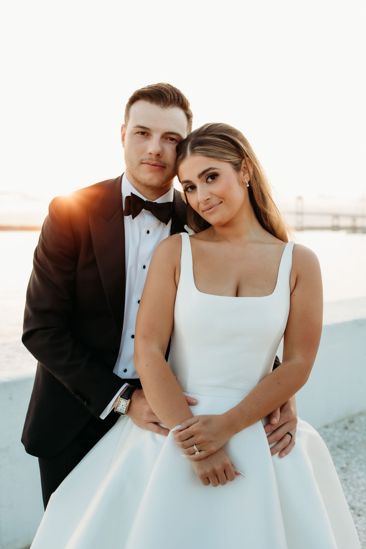A couple in formal wedding attire poses intimately on a beach at sunset.