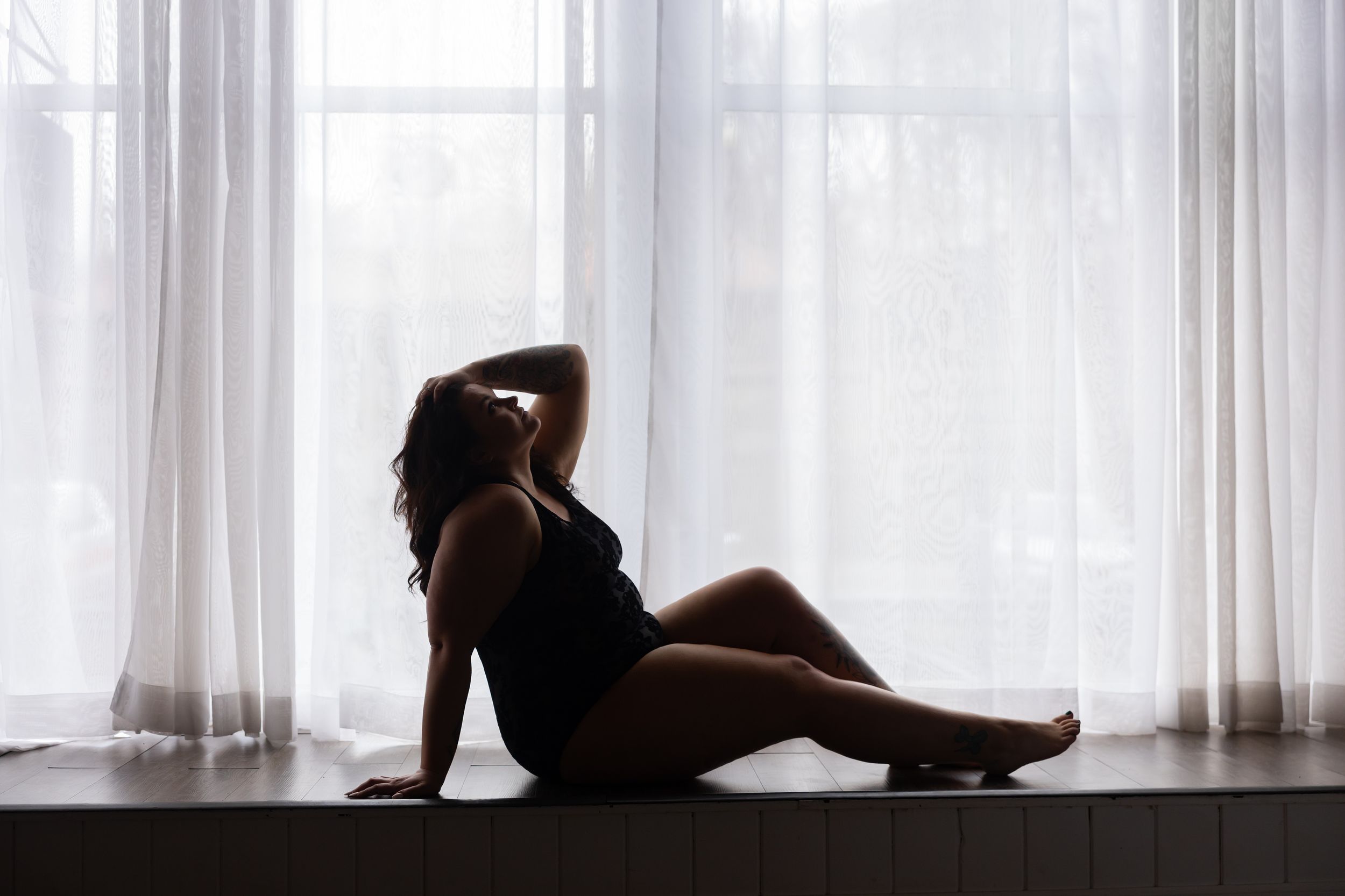 A silhouetted figure sits in contemplative pose by a bright window with sheer curtains.