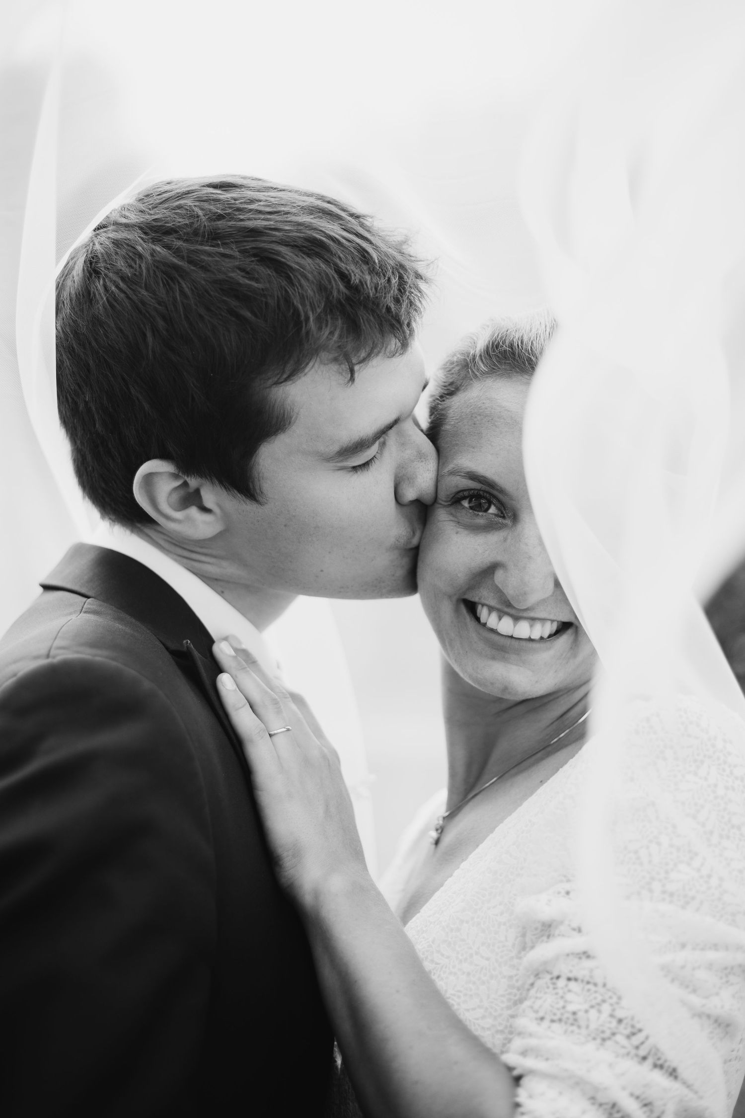 Black and white portrait of groom kissing smiling bride on cheek beneath her flowing wedding veil.