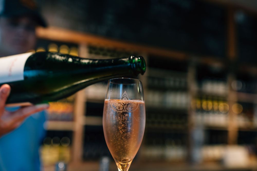 Wine being poured into stemmed glasses on a wooden bar counter.