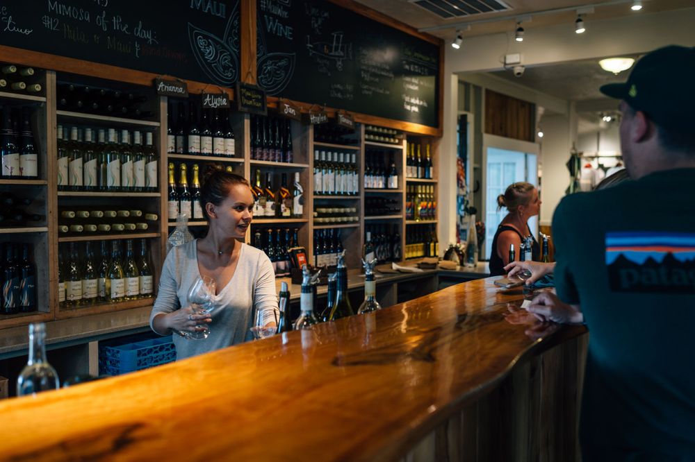 Long wooden bar counter with bottles displayed on shelves and chalkboard menus on the wall.