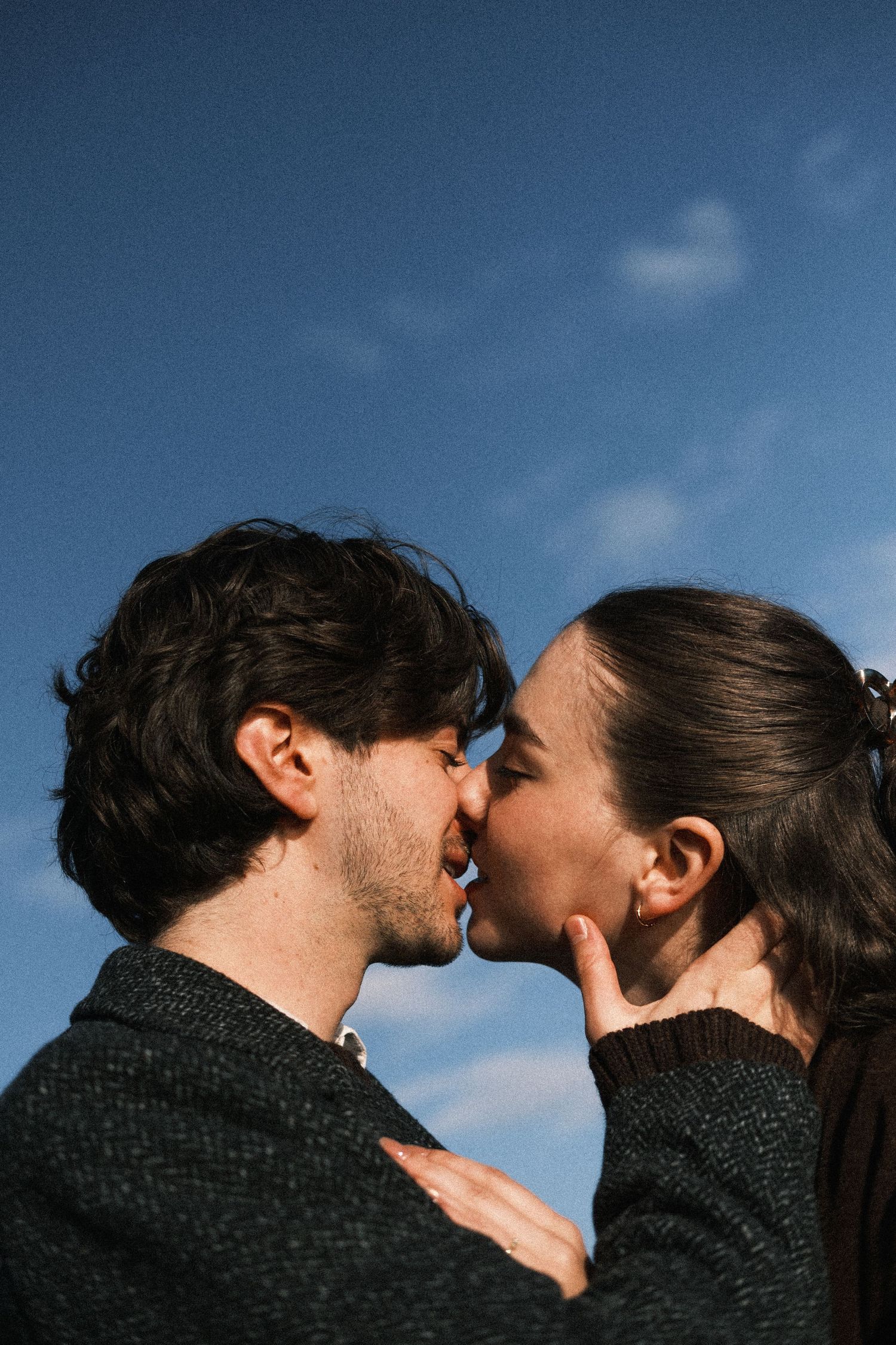 A couple in dark sweaters share a tender kiss against a blue sky background.