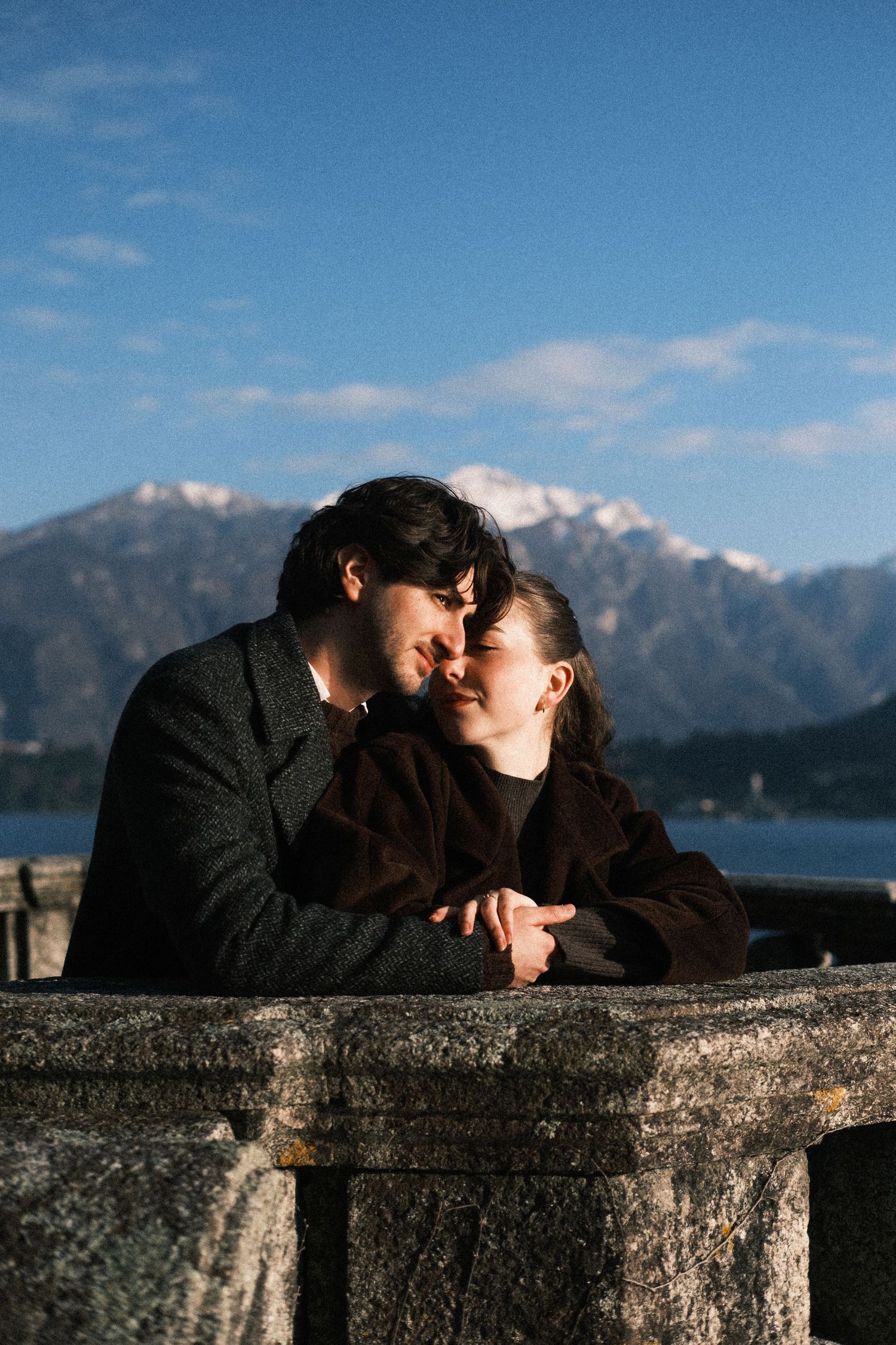 A couple shares an intimate moment while leaning on a stone wall with scenic mountain views and blue skies in the background.
