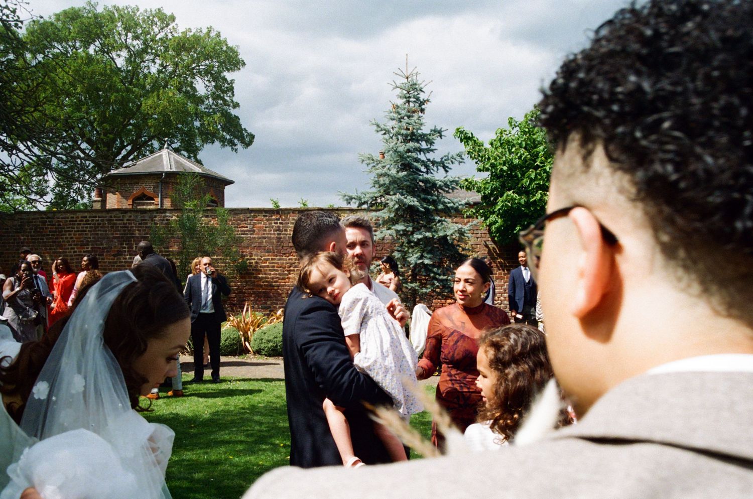 Analogue photo of a toddler resting against father's should whilst looking at bride & groom. Documentary wedding
