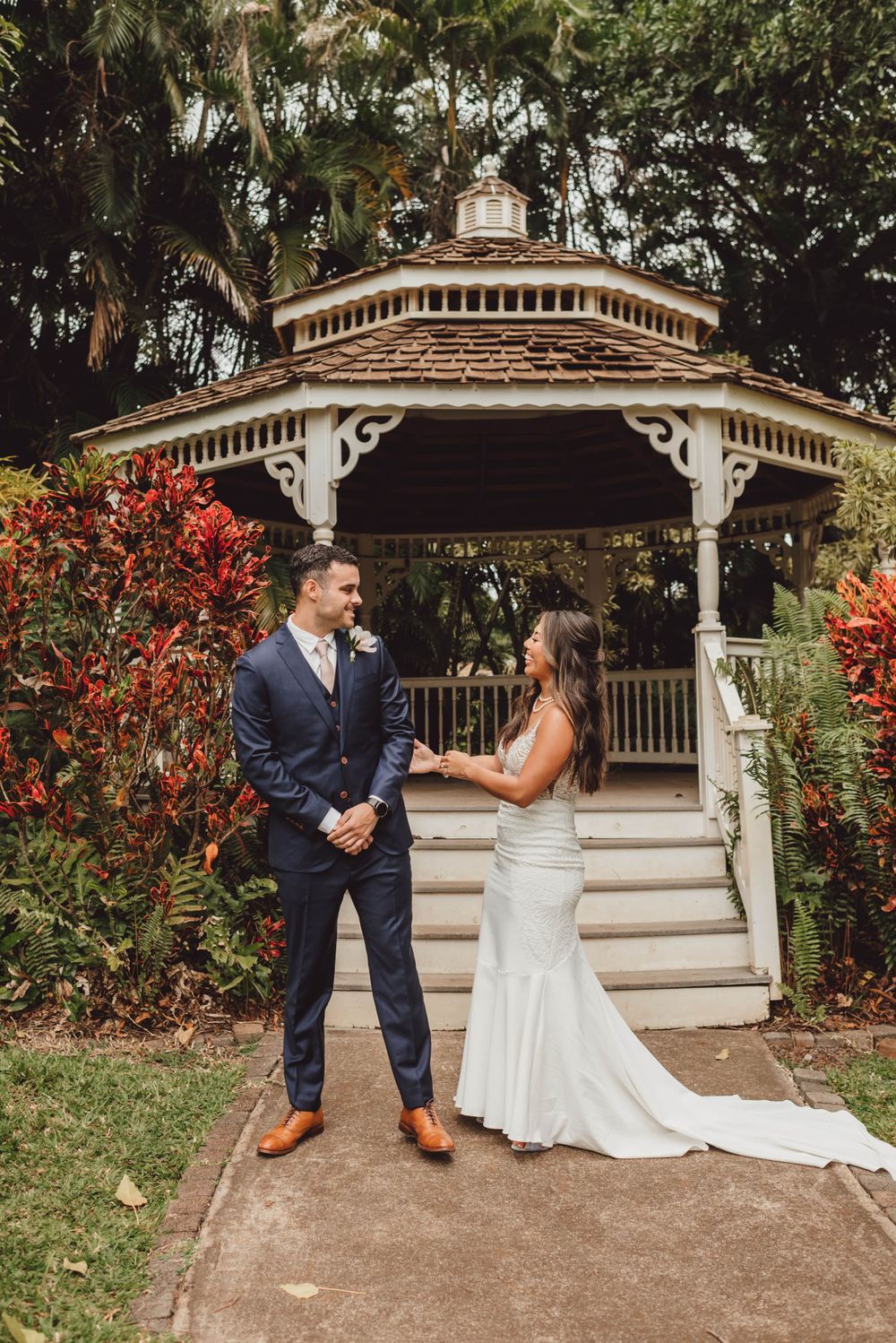 Joyful couple having their first look wedding moment by a decorative white gazebo with vibrant red foliage.