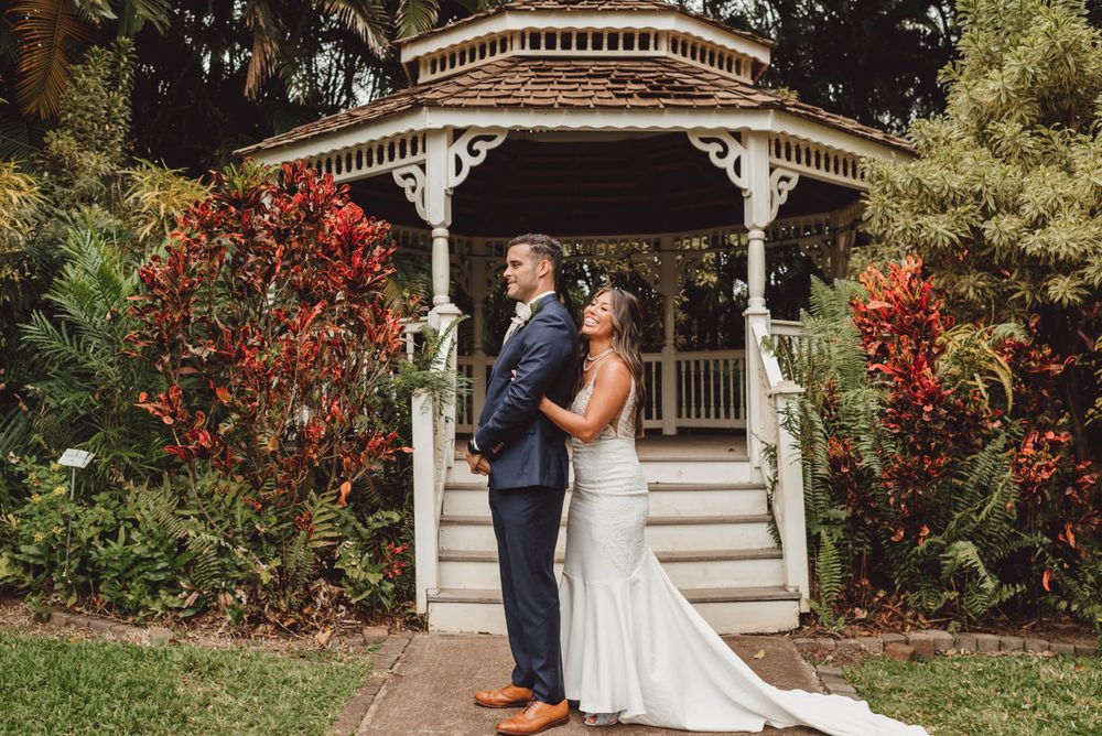A couple shares a sweet first look moment in front of an ornate white gazebo with red flowers blooming nearby.