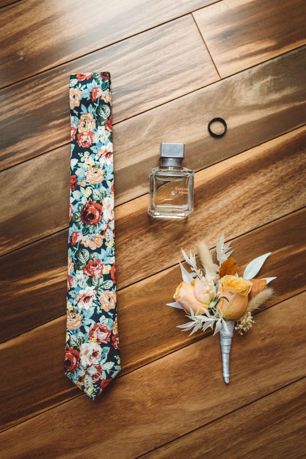 Wedding accessories including floral tie, perfume bottle and boutonniere on wooden floor.