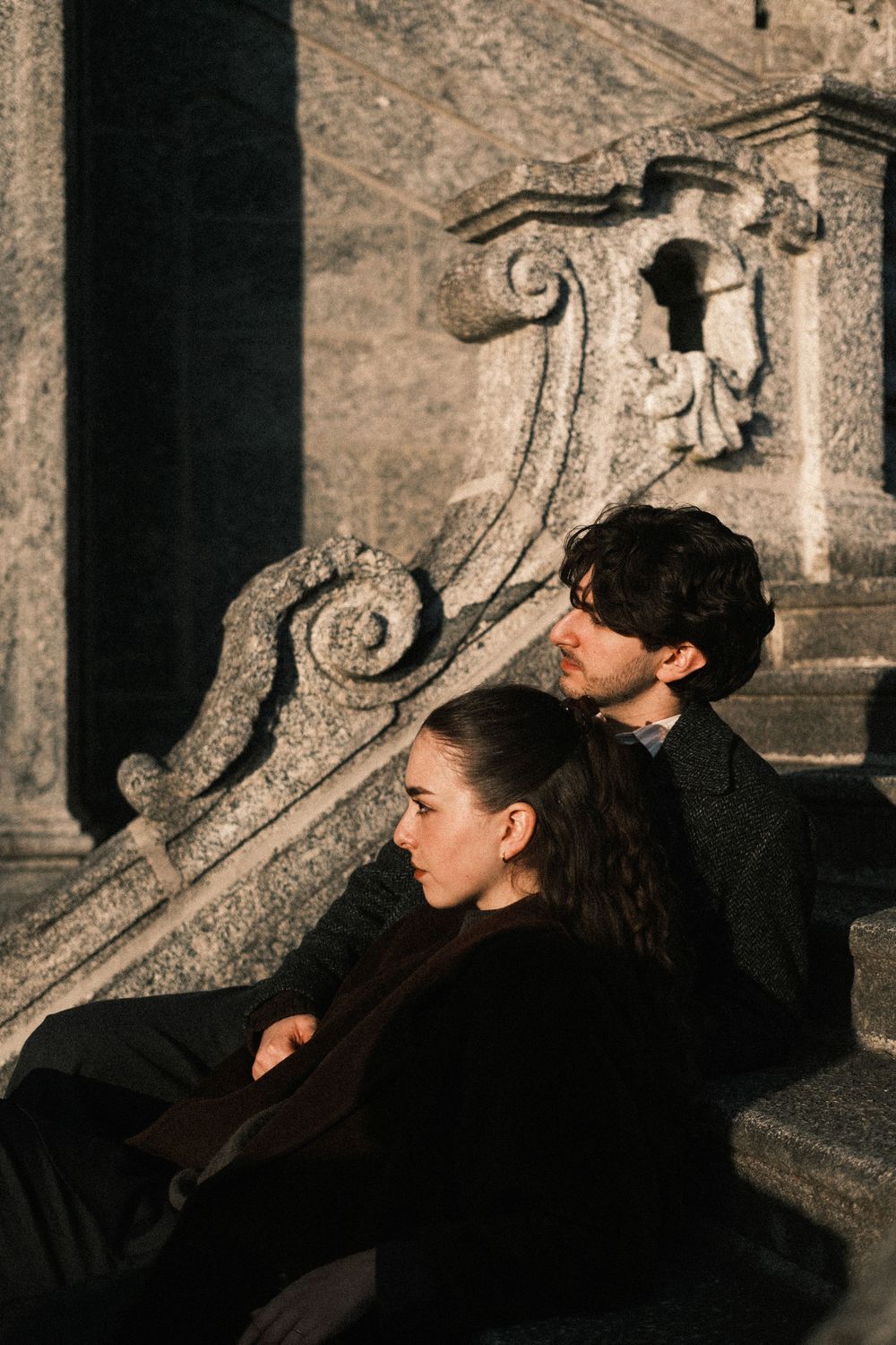 Two people in dark clothing rest against ornate stone architecture with carved scrollwork details in dramatic lighting.