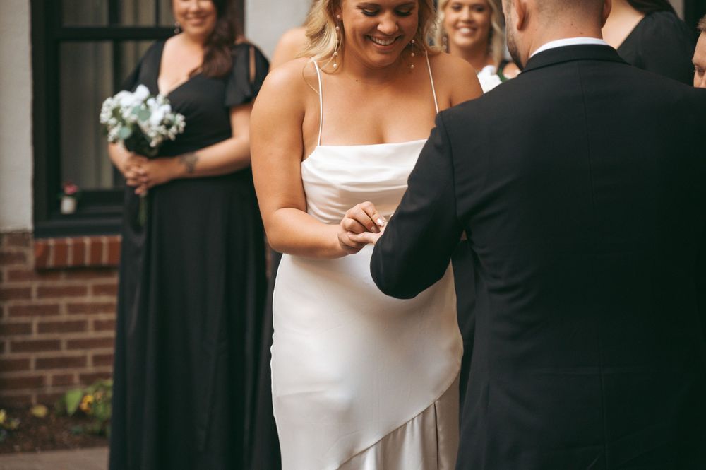 A close-up moment during a wedding ceremony showing hands exchanging rings with bridesmaids in dark dresses in background.