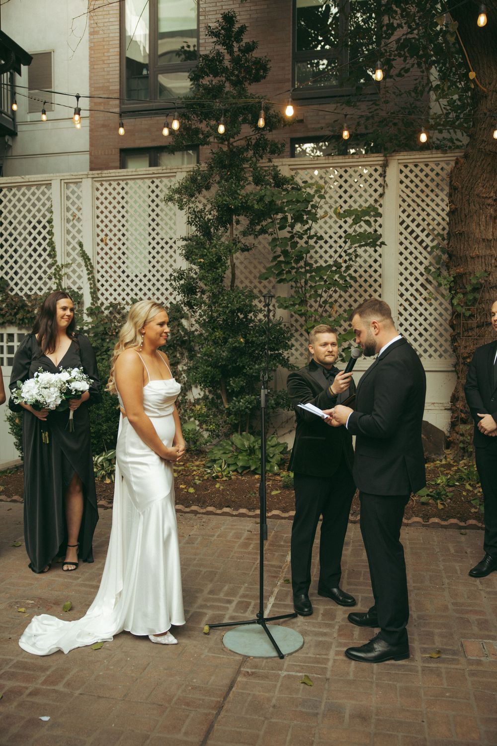A wedding ceremony takes place in a courtyard with white lattice fencing and string lights creating an intimate atmosphere.