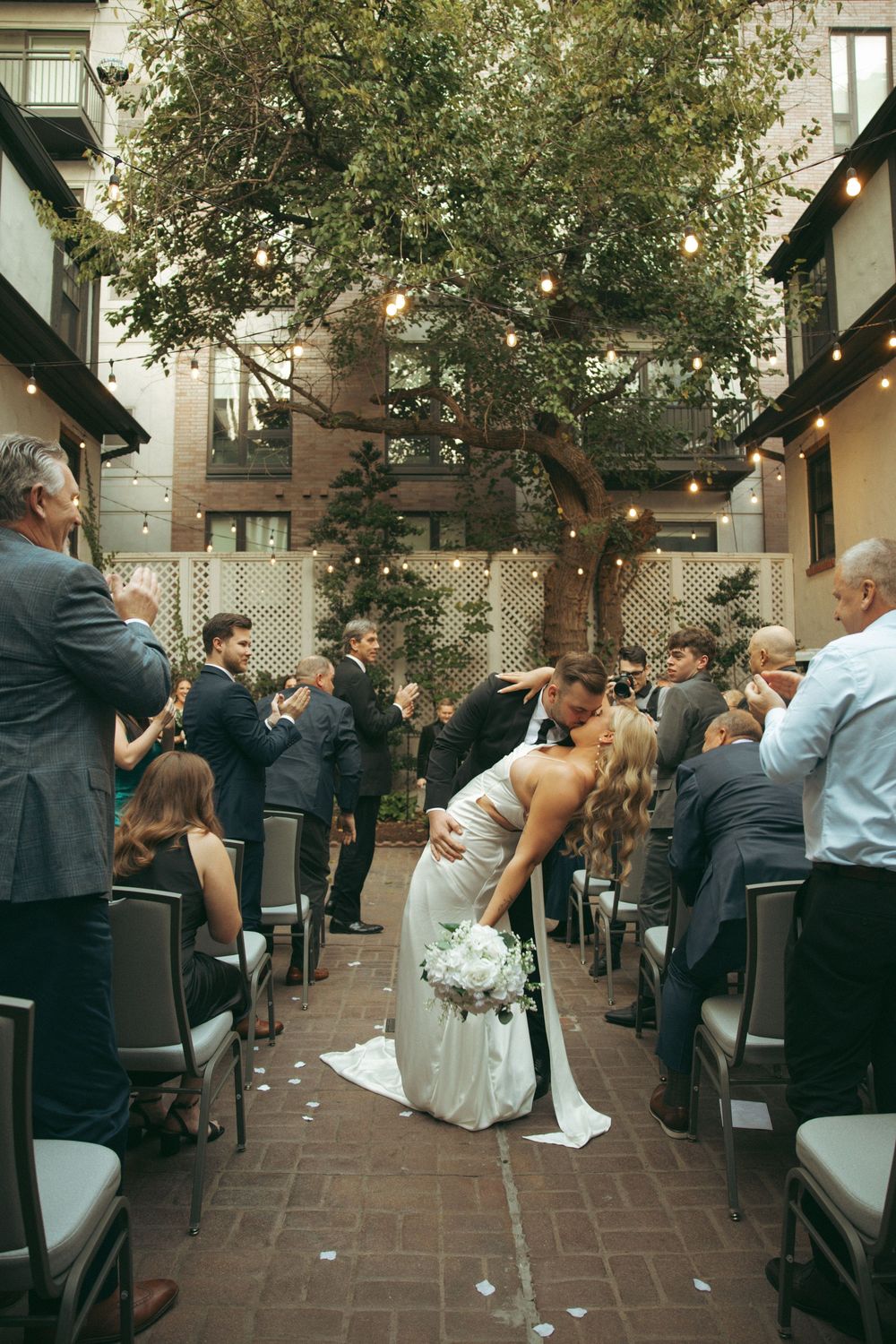 A romantic dip kiss at an outdoor courtyard wedding ceremony with guests watching under a large tree.