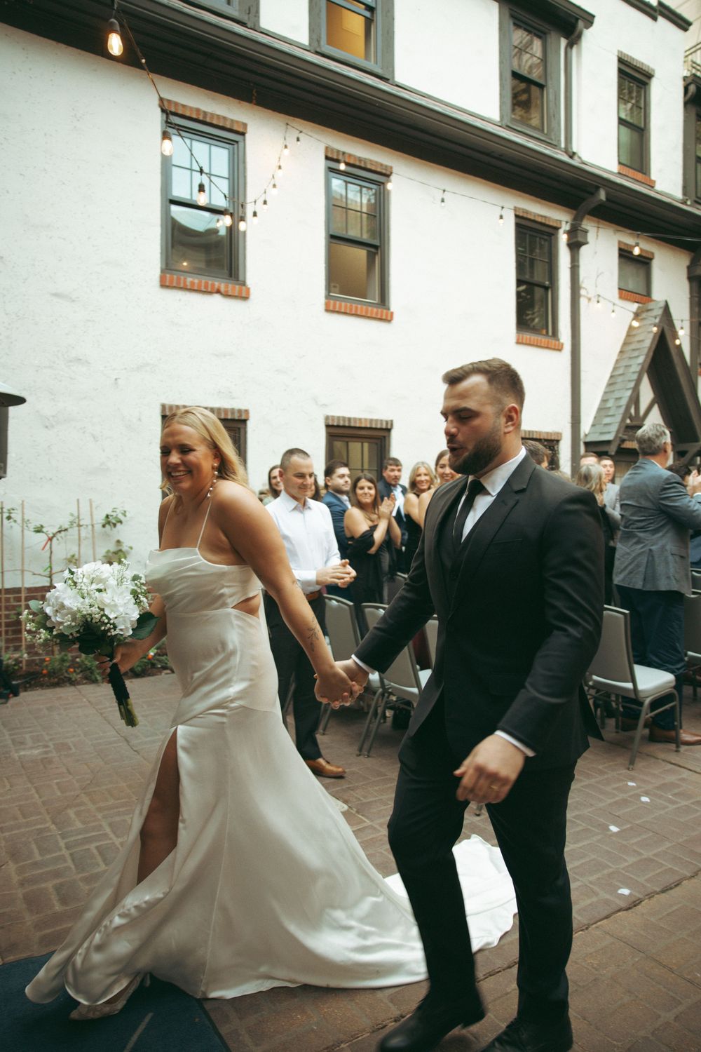 A couple dancing together at an outdoor reception venue with brick pavers and white walls in the background.