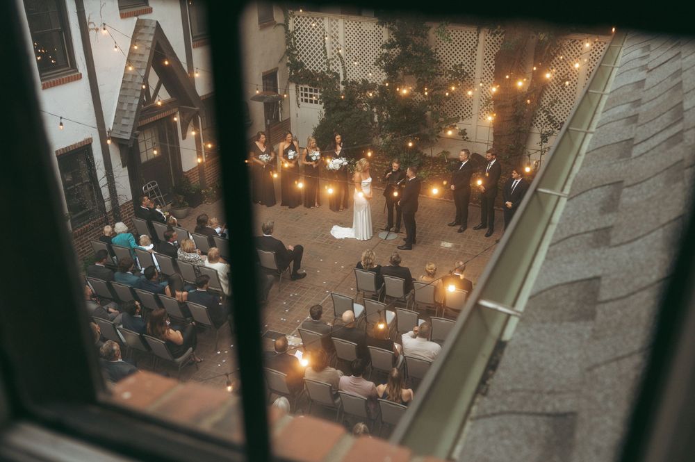 An outdoor evening wedding ceremony viewed through a window, with string lights illuminating guests seated in rows.