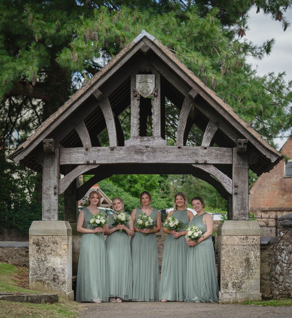 Bridesmaids in sage green dresses pose under a rustic stone archway in a garden setting.