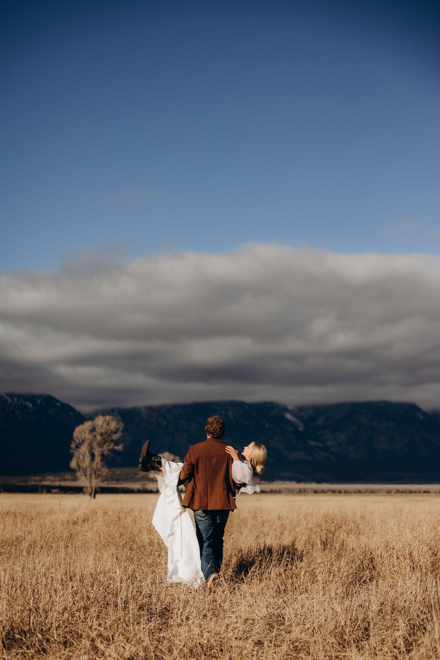 Couple walking through golden wheat field with dramatic mountain backdrop and moody cloudy sky.