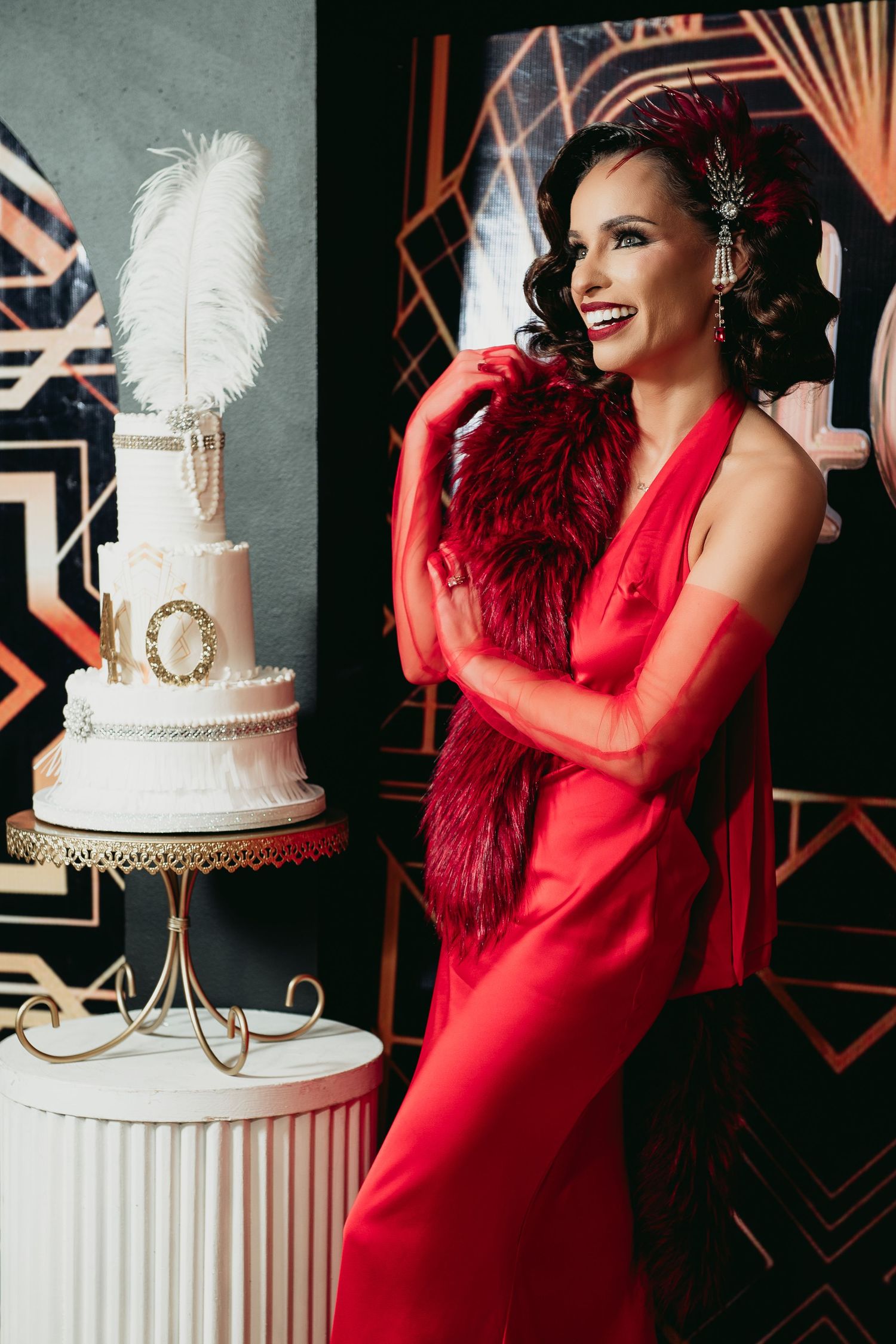 Art deco themed wedding setup with elegant white tiered cake and model in red dress posing against geometric backdrop.