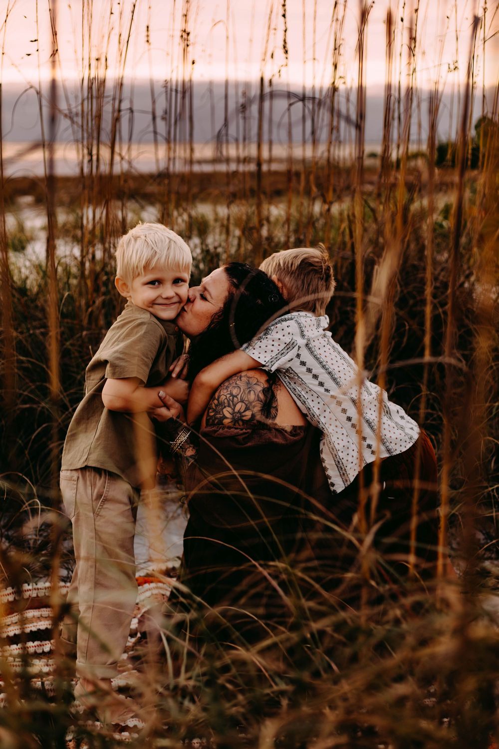 A tender family moment captured through tall grass at sunset in warm golden light.