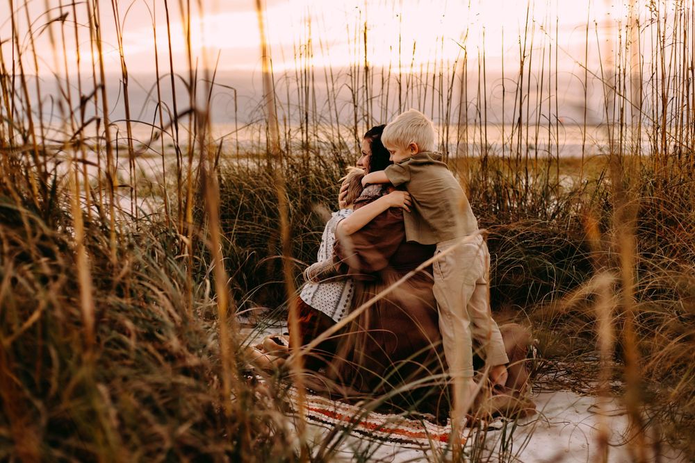 A person in light colored clothing walks through tall reeds and marsh grass at sunset near a body of water.