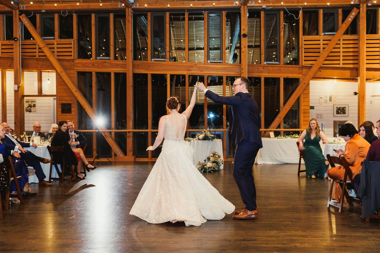 Dancing couple twirls on wooden floor during wedding reception in rustic barn setting.