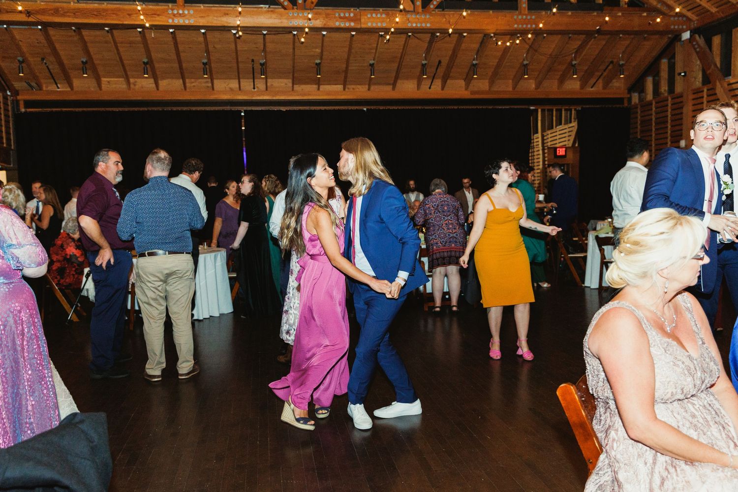 Wedding guests dancing and celebrating on wooden dance floor under string lights.