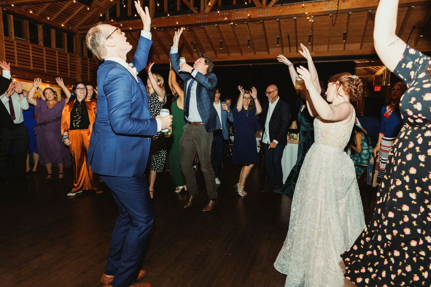 Energetic wedding reception dance party with guests raising hands under rustic wooden beams.