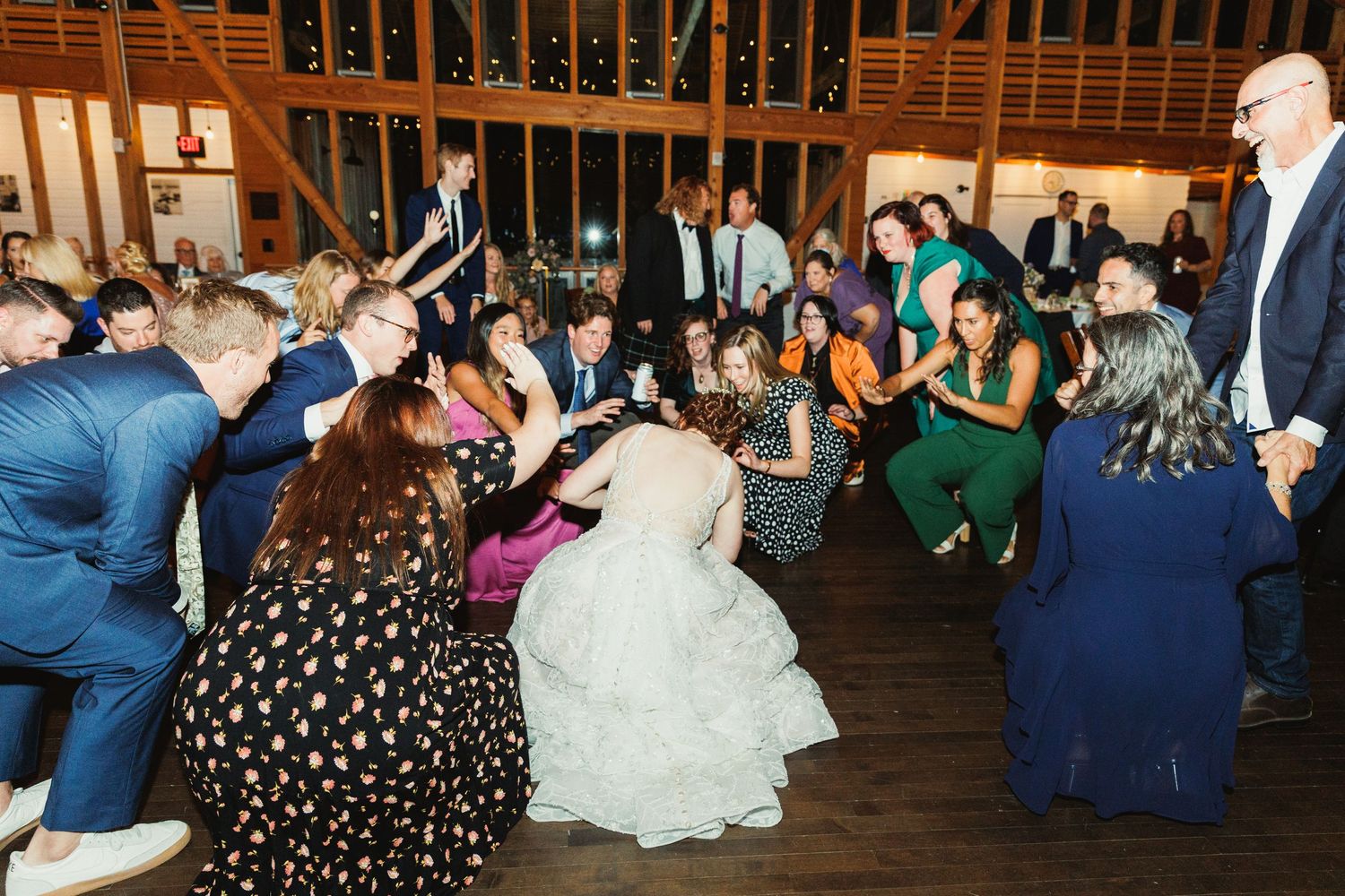 Wedding guests dancing enthusiastically on a wooden dance floor under wooden beams and string lights.