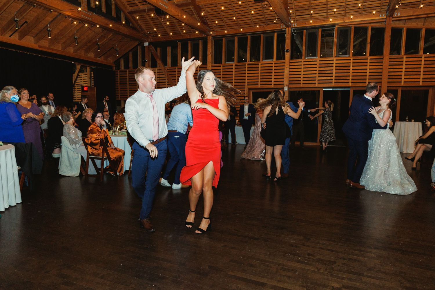 Energetic dance party at a wedding reception with string lights illuminating the wooden barn venue.