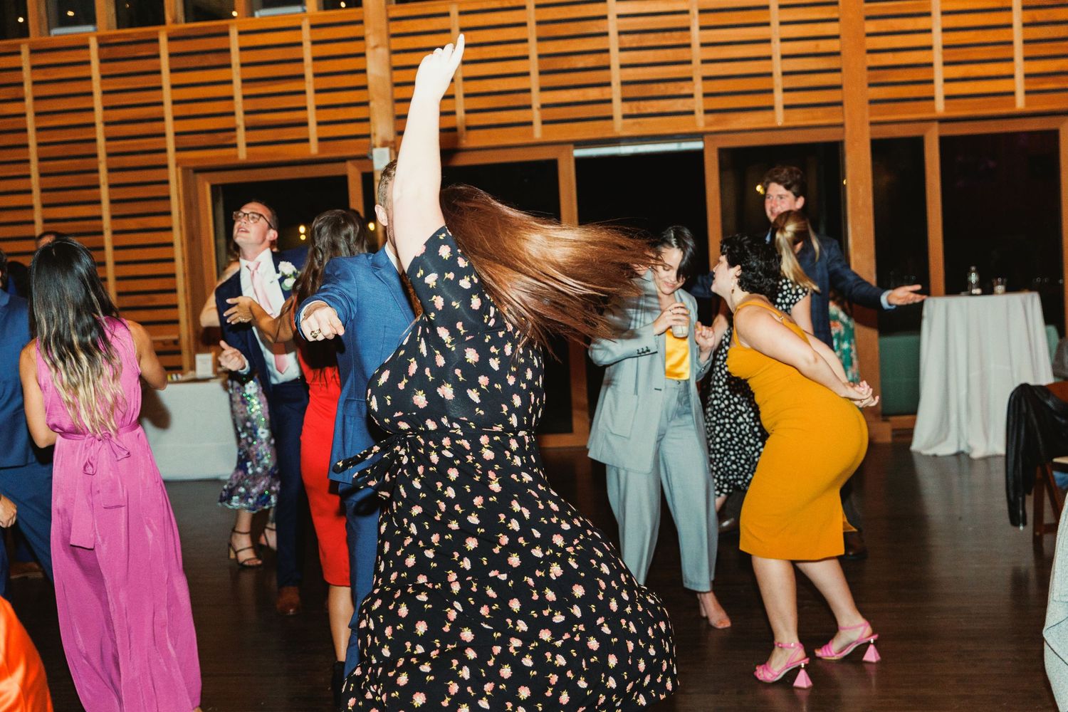 Wedding guests dancing joyfully in colorful dresses under string lights in a barn venue.