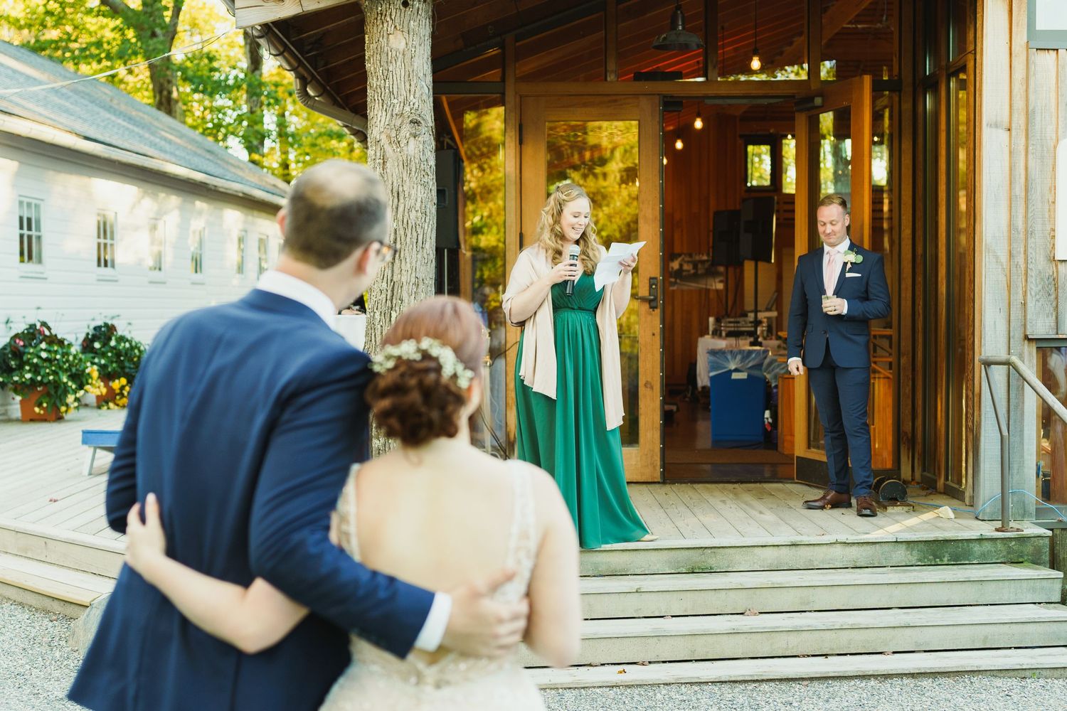 Wedding guests in formal attire gather at the entrance of a rustic venue with wooden doors and greenery.
