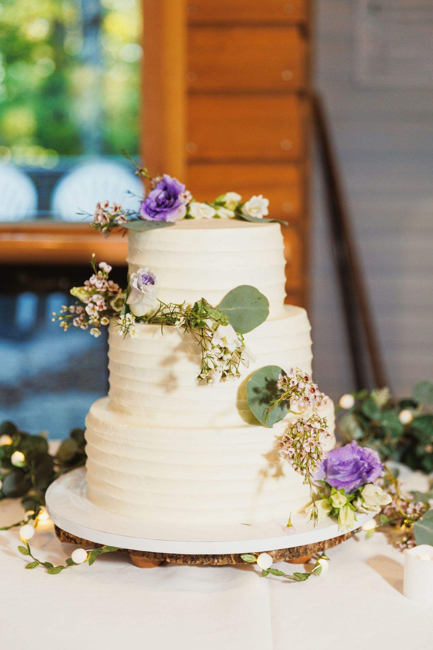 Three-tiered white wedding cake decorated with purple flowers, eucalyptus leaves and delicate floral accents.