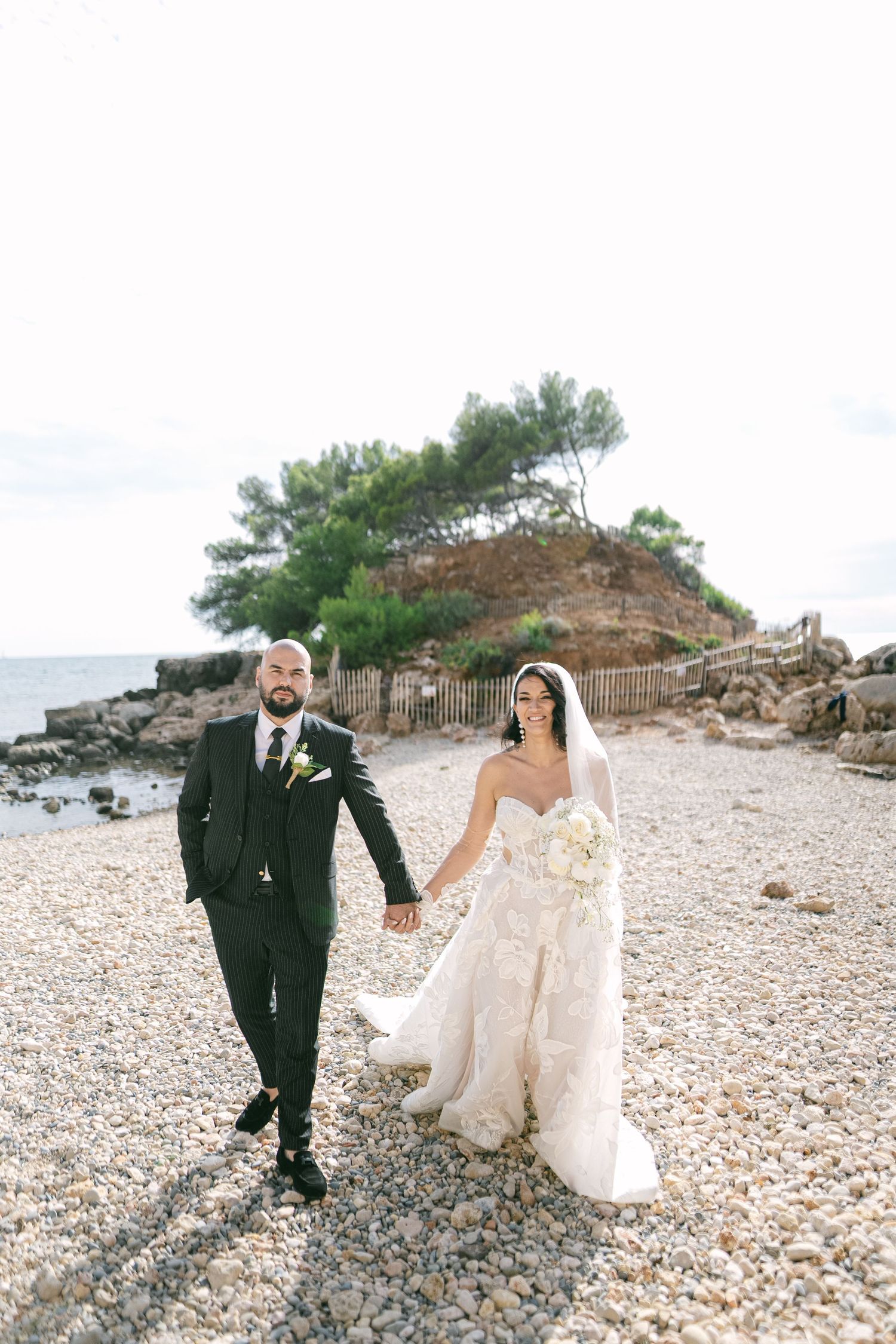 A couple in wedding attire walks together on a rocky beach with trees and ocean in the background.