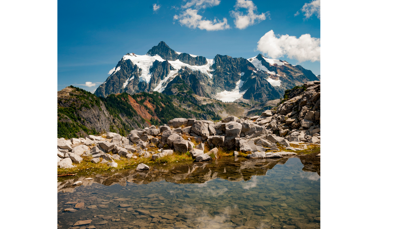 Snowy Mt. Baker peaks reflect in a clear alpine lake surrounded by rocky terrain under bright blue skies at Artist Point