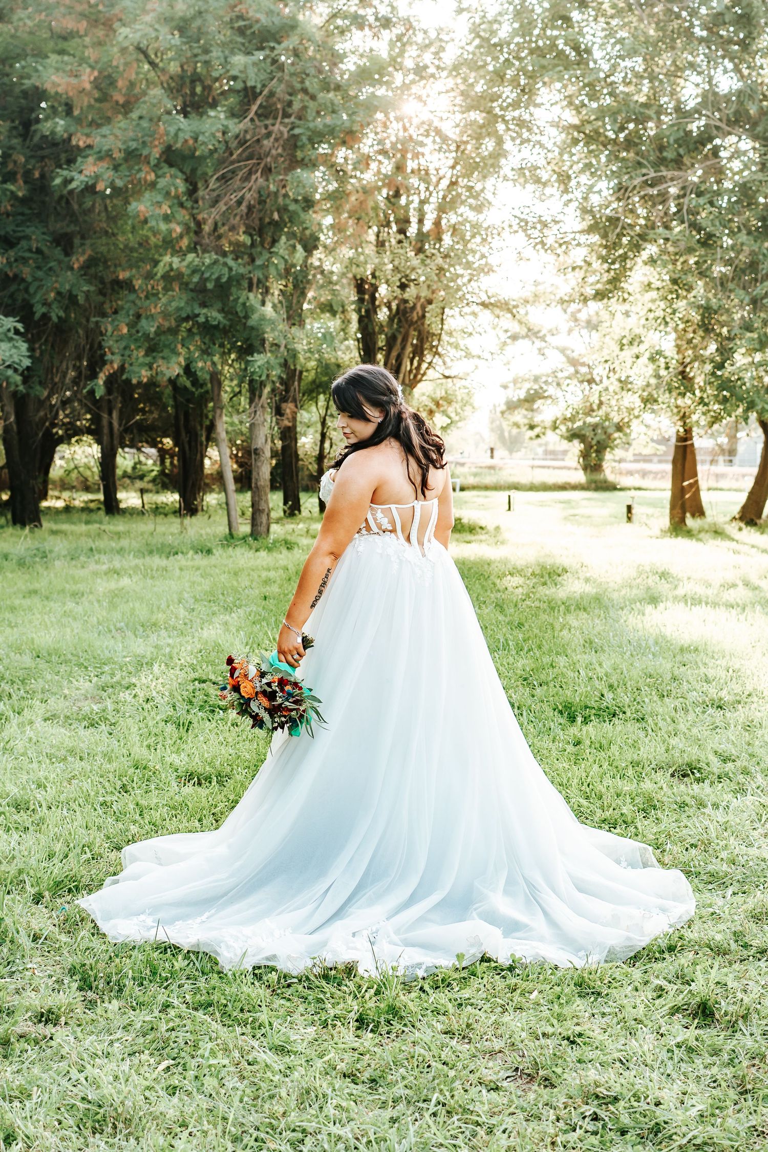 A bride in a flowing white ballgown holds a colorful bouquet while standing in a sunlit pasture in Irrigon, Oregon.