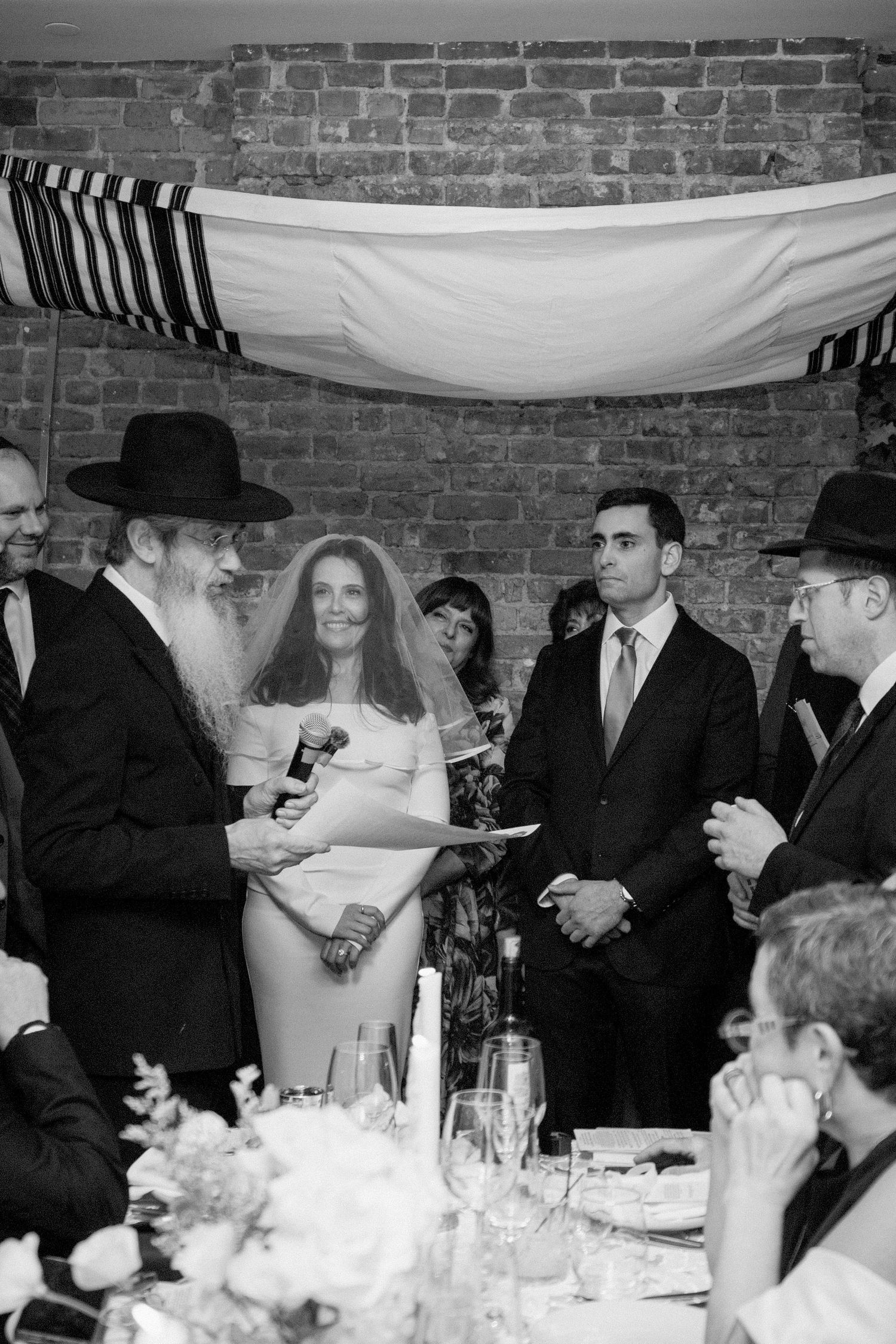 Wedding reception scene with guests seated at tables under draped fabric in brick-walled venue.