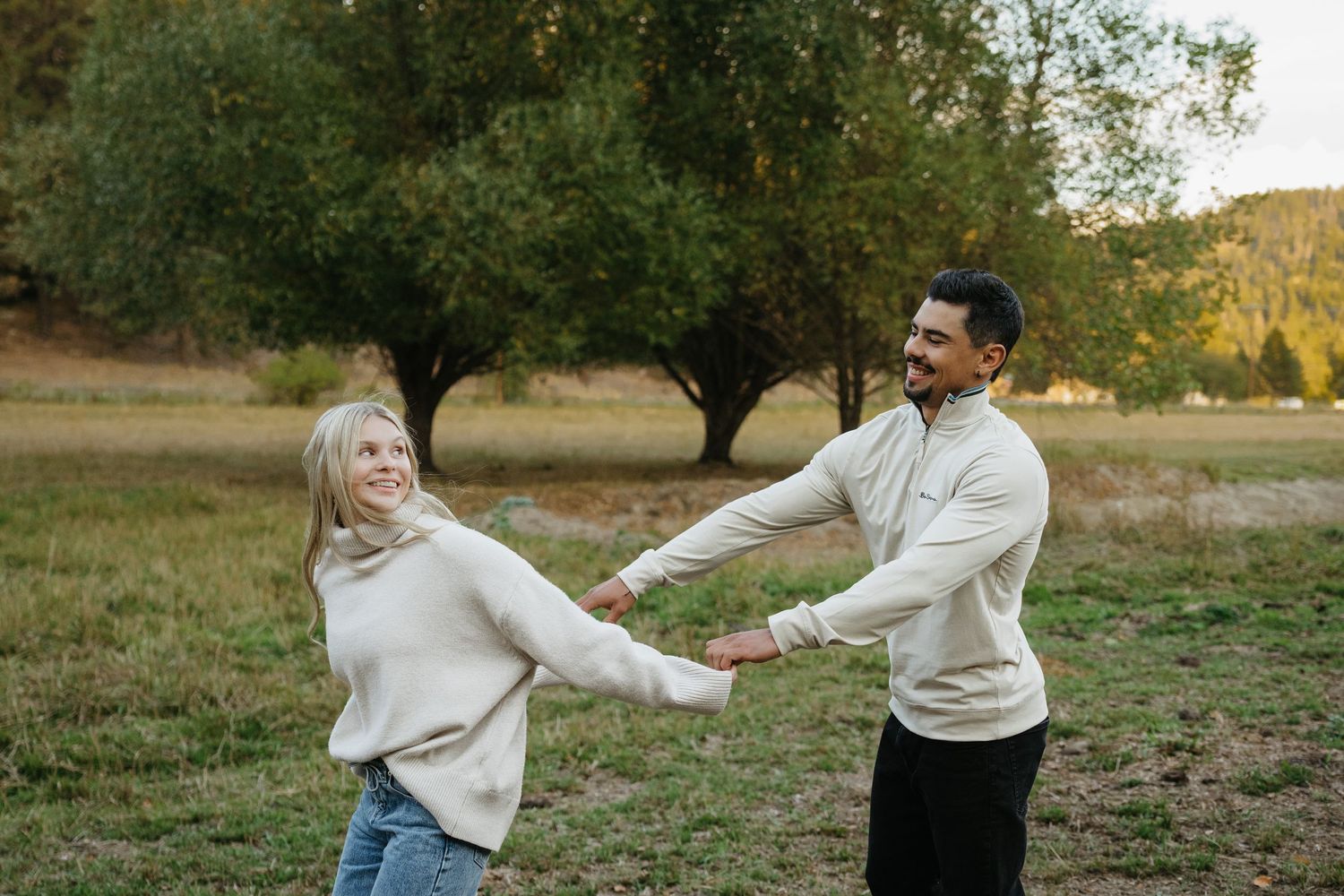A couple holds hands while dancing and laughing together in a grassy field with trees at sunset.