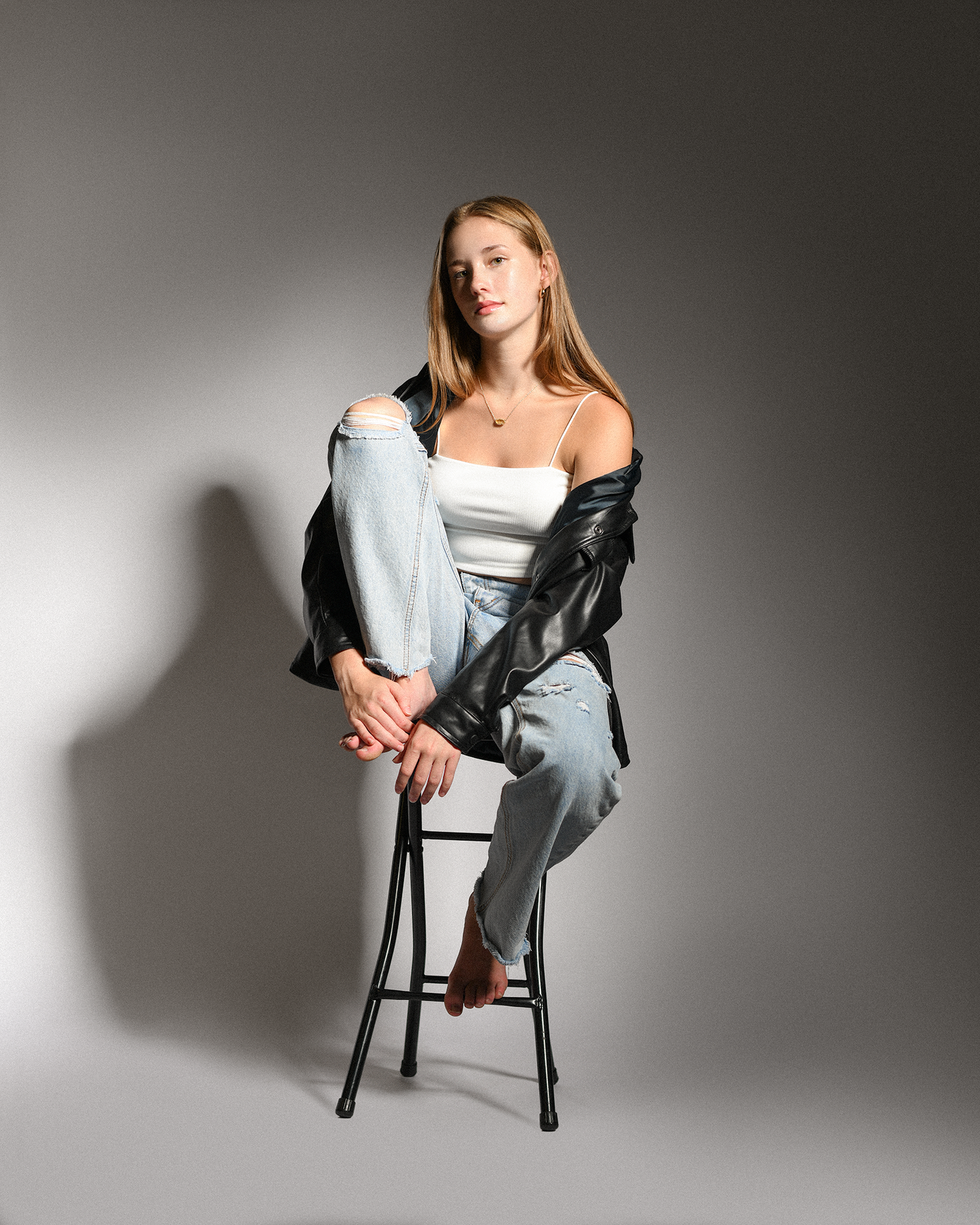 Young woman in ripped jeans and leather jacket sitting on a stool posing in a studio with gray background.