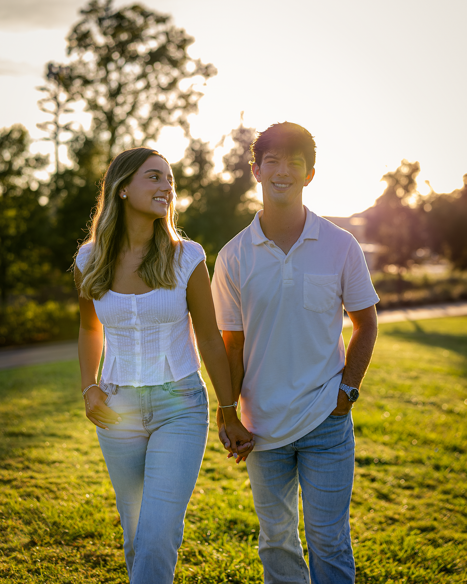 Couple holding hands walking through a sunny park at golden hour, wearing white tops and jeans.