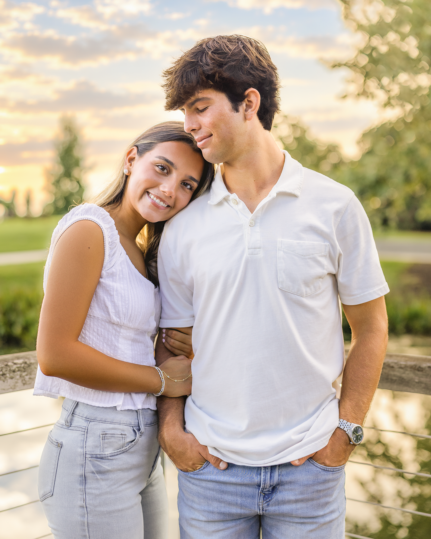 A young couple in white tops and jeans pose together outdoors at sunset, smiling warmly in a park setting.