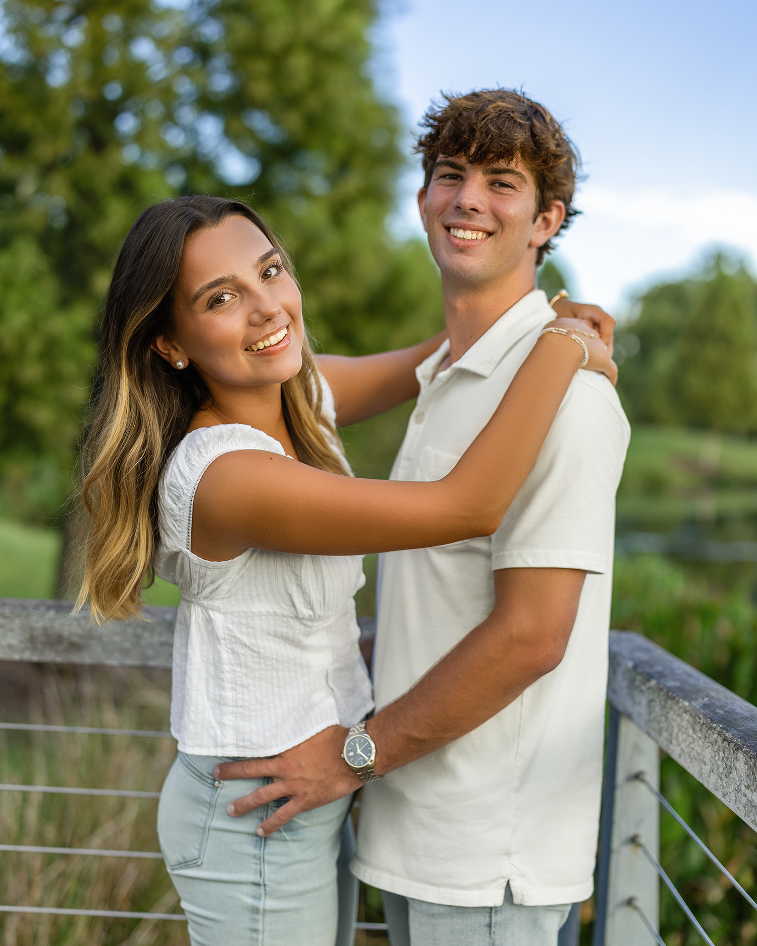 A smiling young couple poses outdoors on a bridge, wearing white outfits, surrounded by lush green trees.