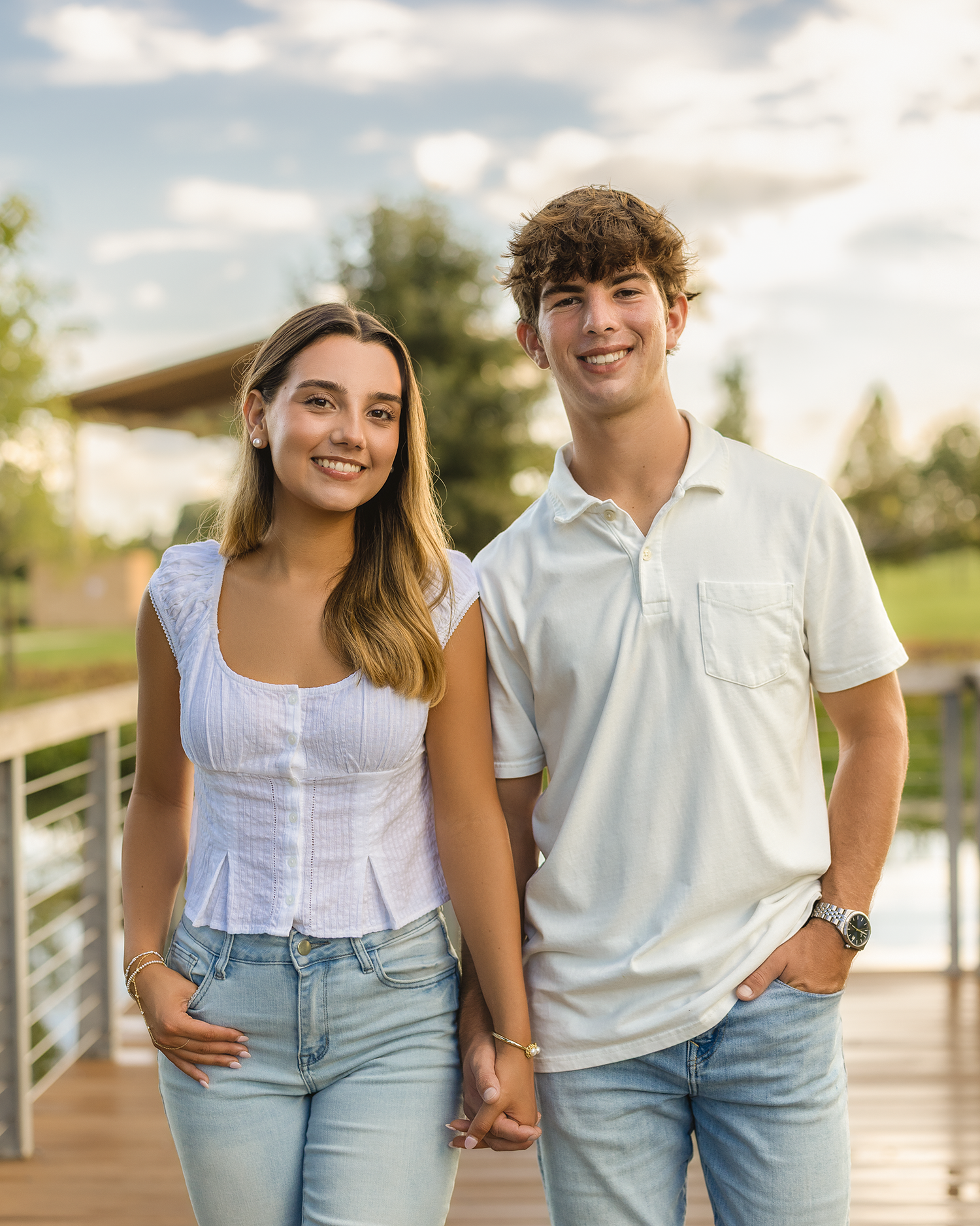Young couple holding hands and smiling outdoors on a wooden deck with a scenic park background.
