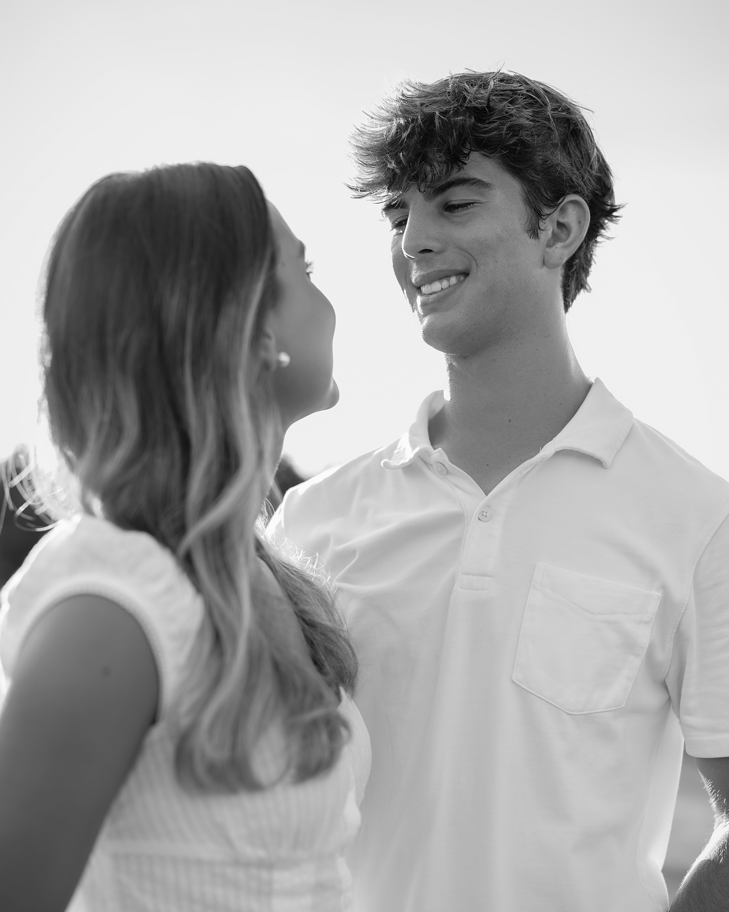 Black and white photo of a smiling teenage boy in a white polo shirt looking at a girl with long hair.