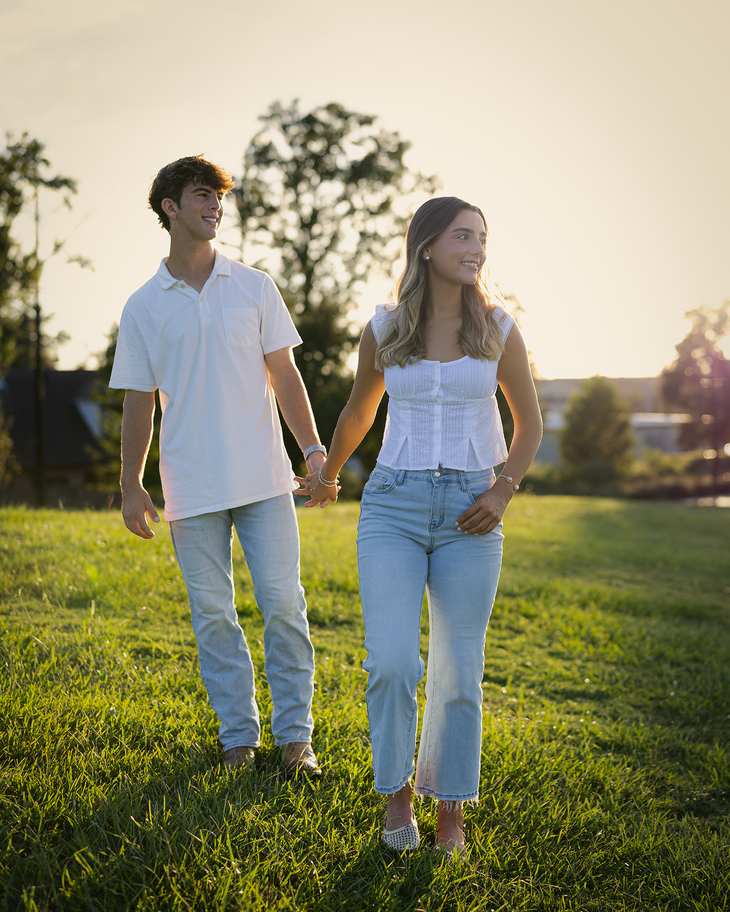 Young couple holding hands walking on green grass field at golden hour, wearing matching white tops and light jeans.