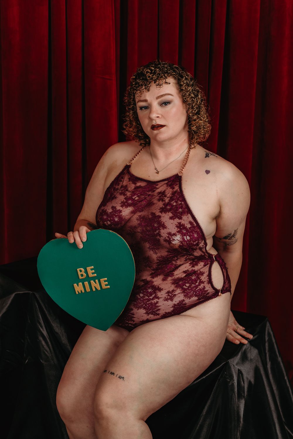 A boudoir photoshoot with burgundy lace and a green heart-shaped prop against a dark red curtain background.