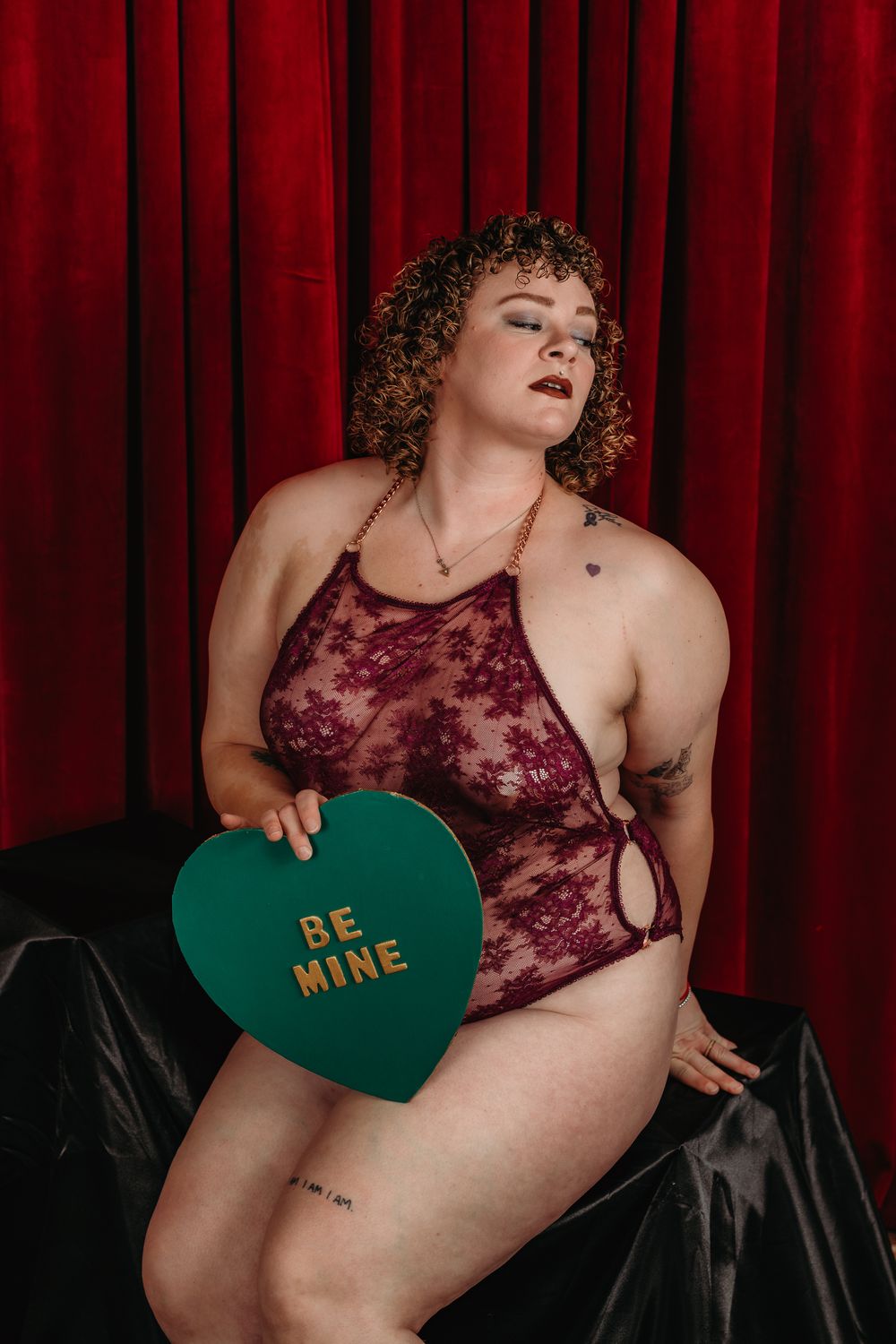 A burgundy lace boudoir photoshoot with a green heart-shaped prop against a red curtain backdrop.