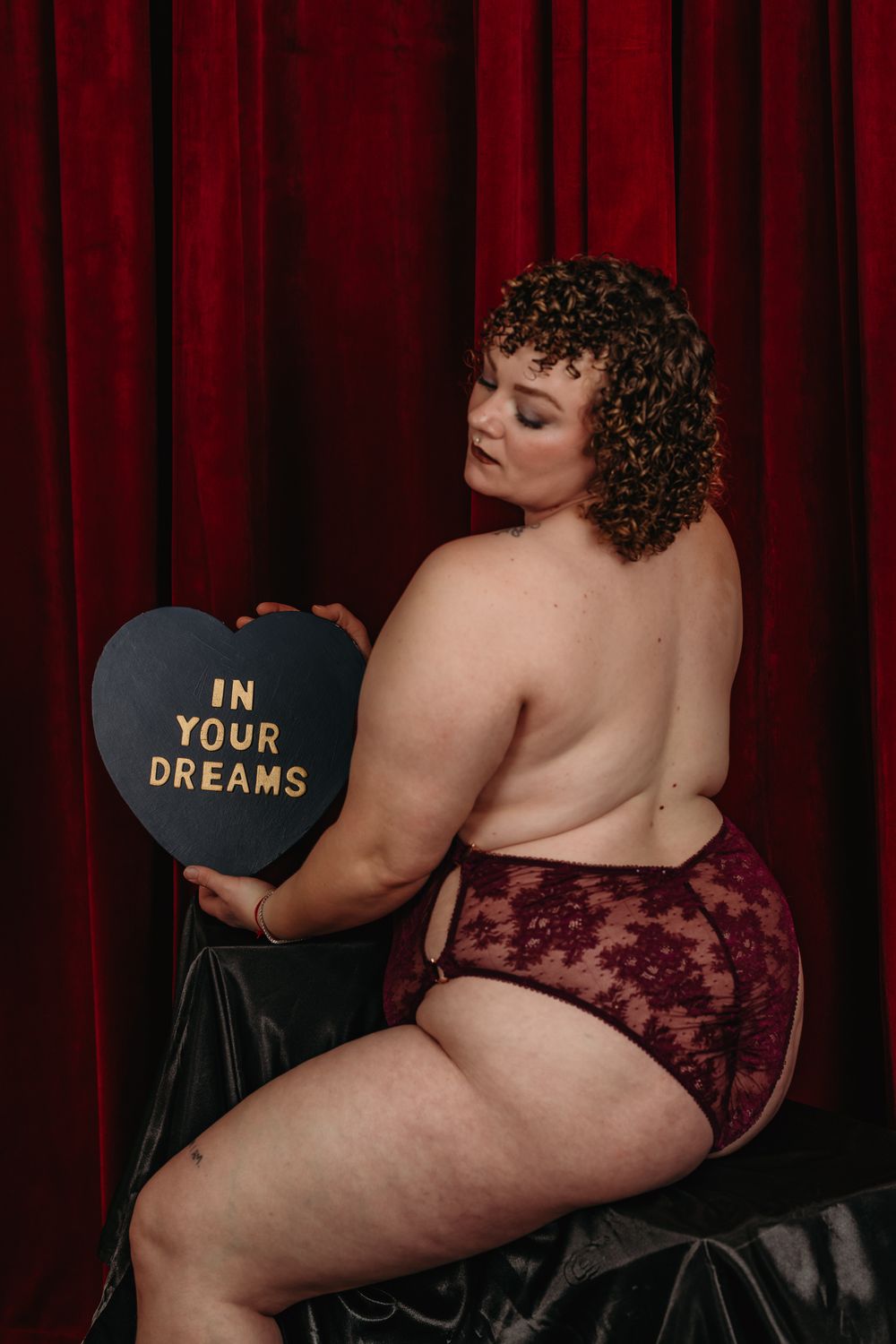 Boudoir photo featuring curly-haired model in burgundy lace lingerie holding black heart pillow against red curtain backdrop.