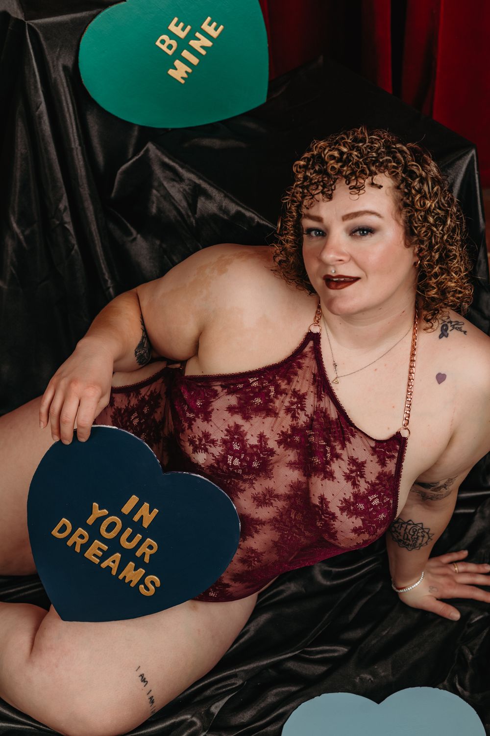 A boudoir photo series showing a person in burgundy lace lingerie posing with decorative heart pillows on dark fabric.