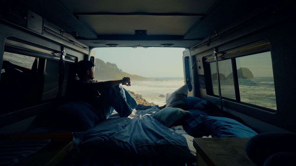 Interior view of van bed setup with coastal scenery visible through back window.